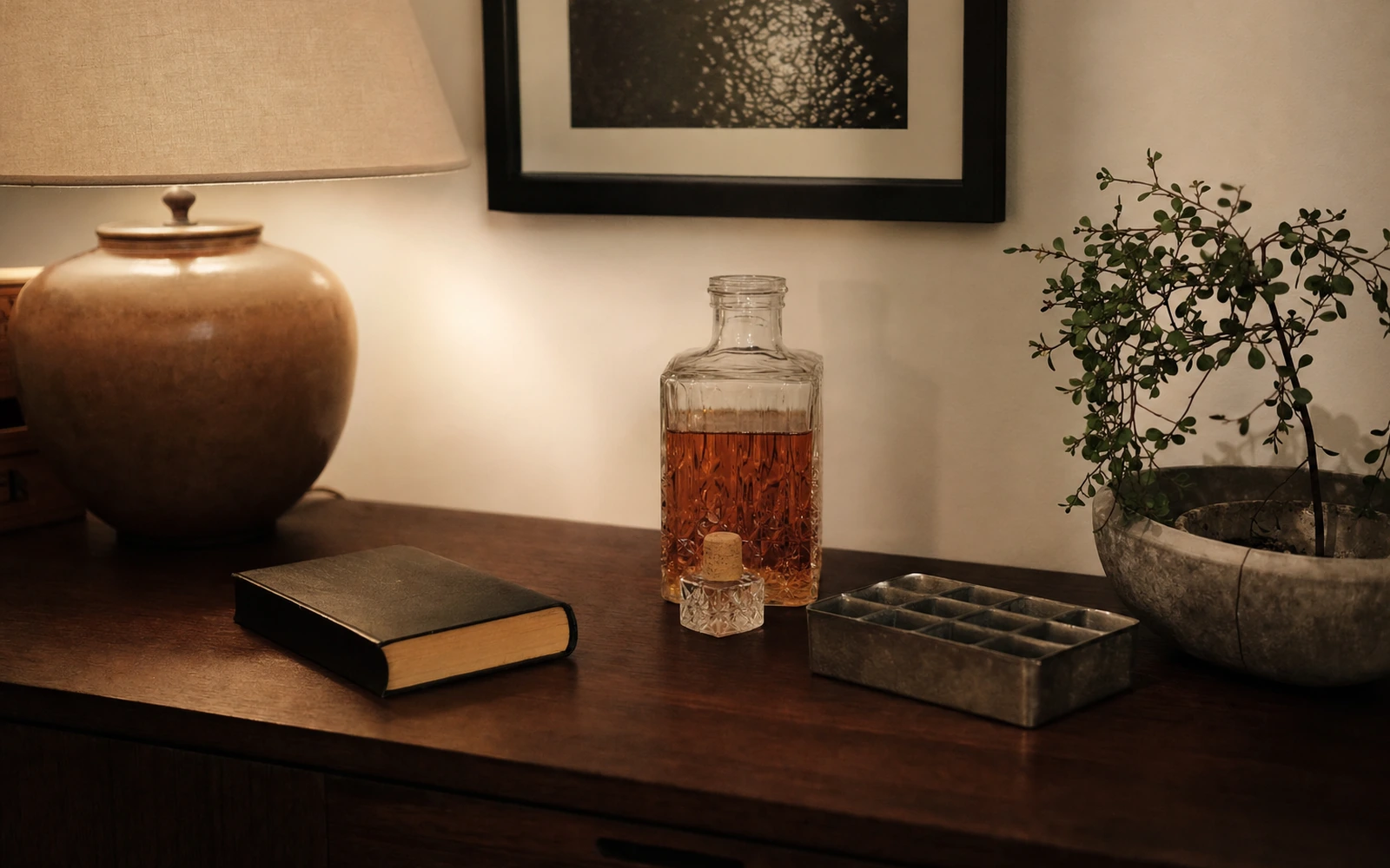 Warm home shelf with unlabeled whiskey decanter, cork, ice tray, and book arranged away from the bedside.