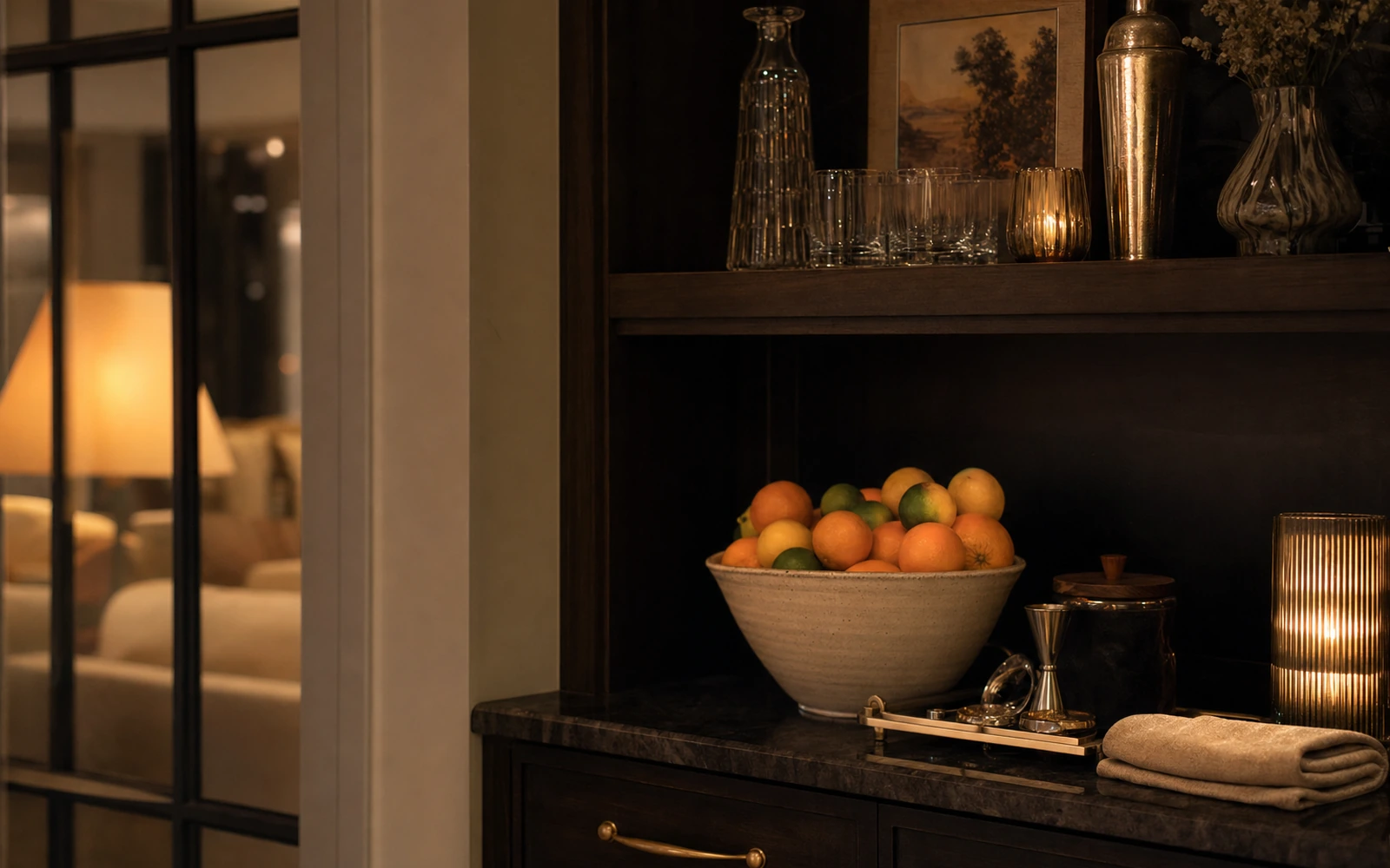 A dark wood bar cabinet at night with a bowl of citrus, glassware, and a candle.