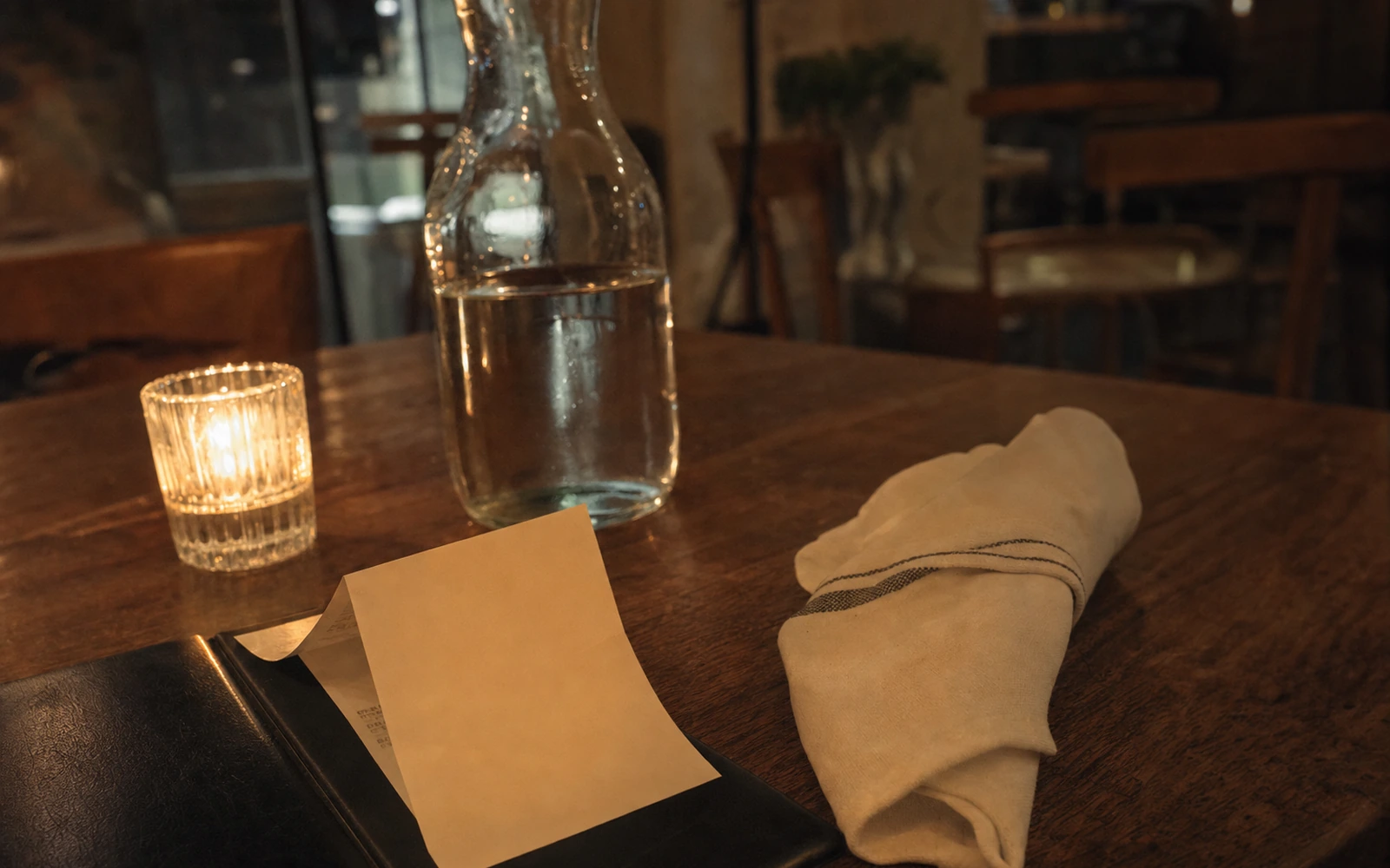 Warm restaurant table with a folded receipt, card, and water carafe, no readable prices or logos.