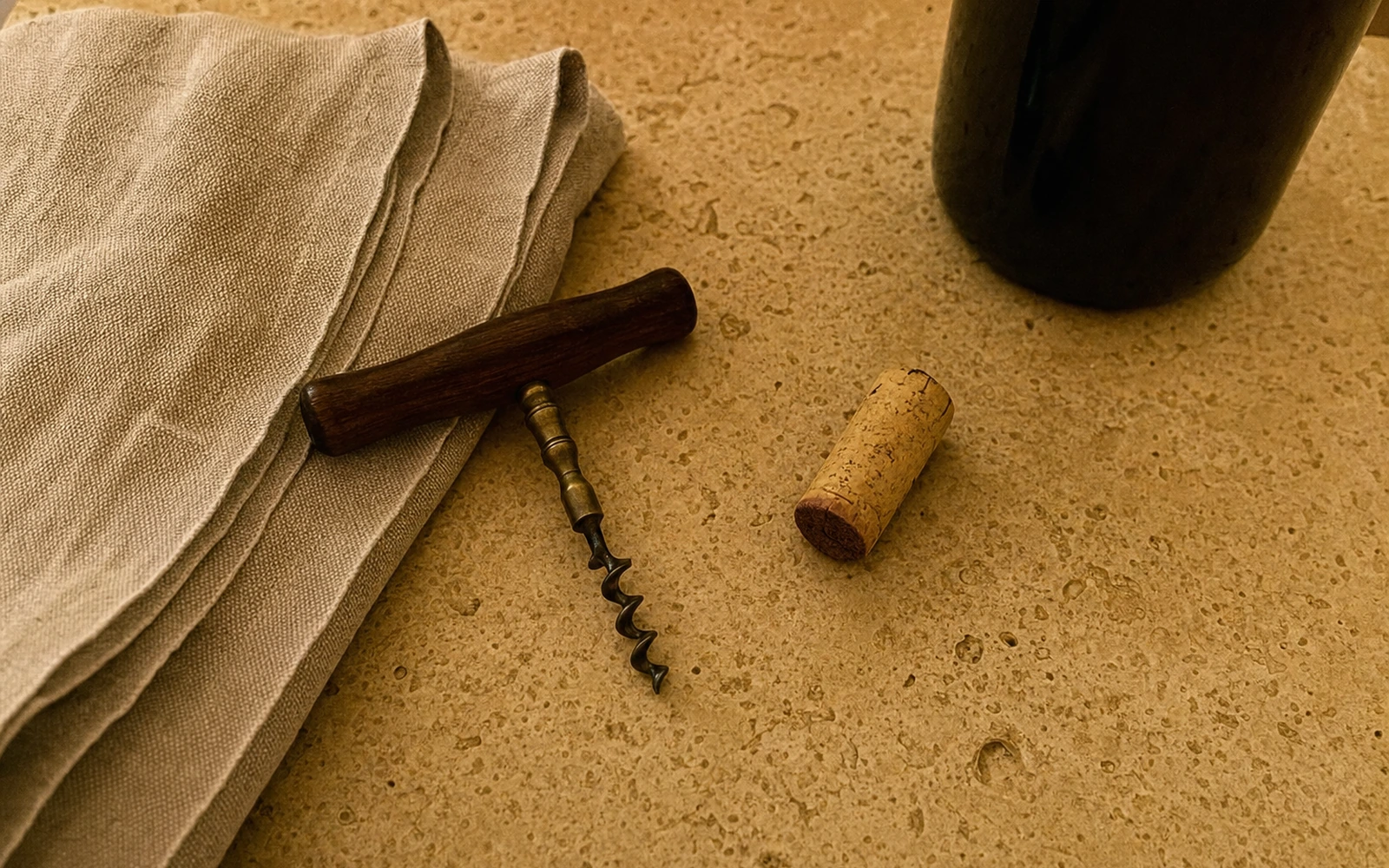 Overhead view of a corkscrew and cork on a stone counter beside a linen towel and a dark wine bottle.